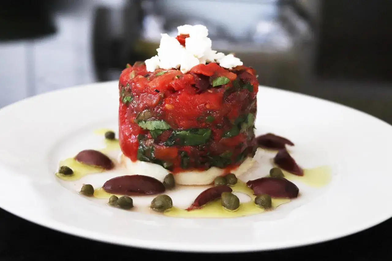 Delicioso tartar de tomate con queso fresco y aceitunas, adornado con alcaparras y aceite de oliva.