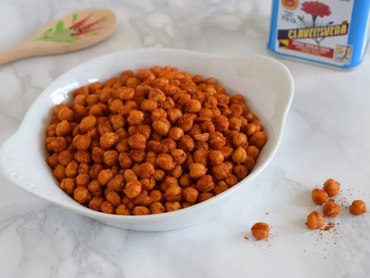 Un tazón blanco lleno de garbanzos fritos crujientes y especiados. Una lata de especias y una cuchara de madera decorada están al fondo.