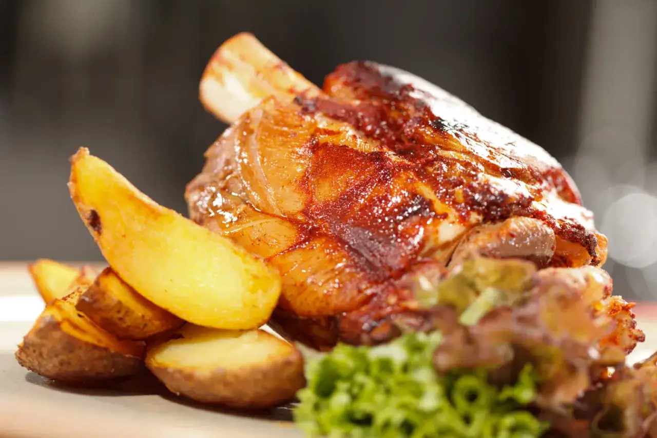 Delicioso codillo de cerdo asado, dorado y jugoso, acompa&ntilde;ado de patatas gajo crujientes y una guarnici&oacute;n verde.