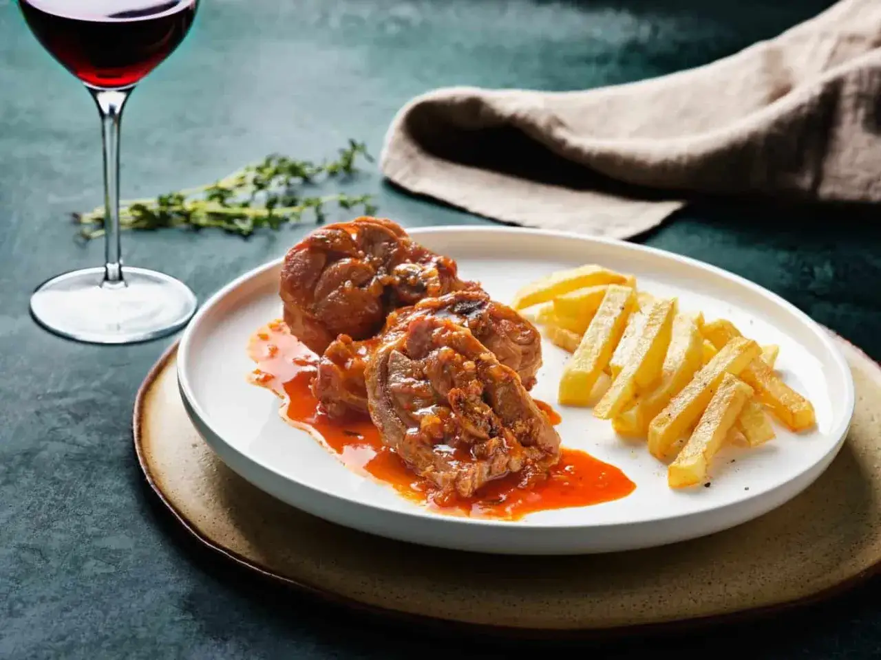 Delicioso cordero al chilindr&oacute;n en un plato blanco, acompa&ntilde;ado de patatas fritas y una copa de vino tinto.