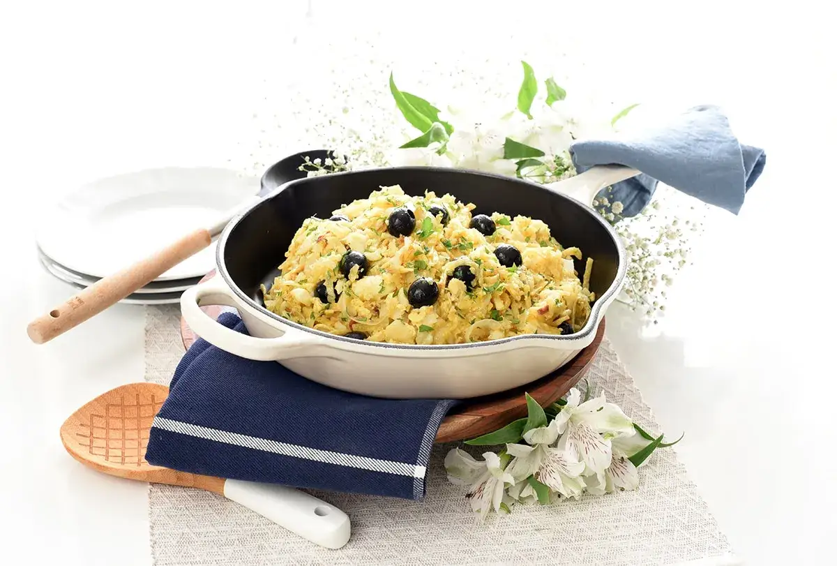 Delicioso bacalao dorado con aceitunas negras y perejil, servido en sartén, listo para disfrutar.