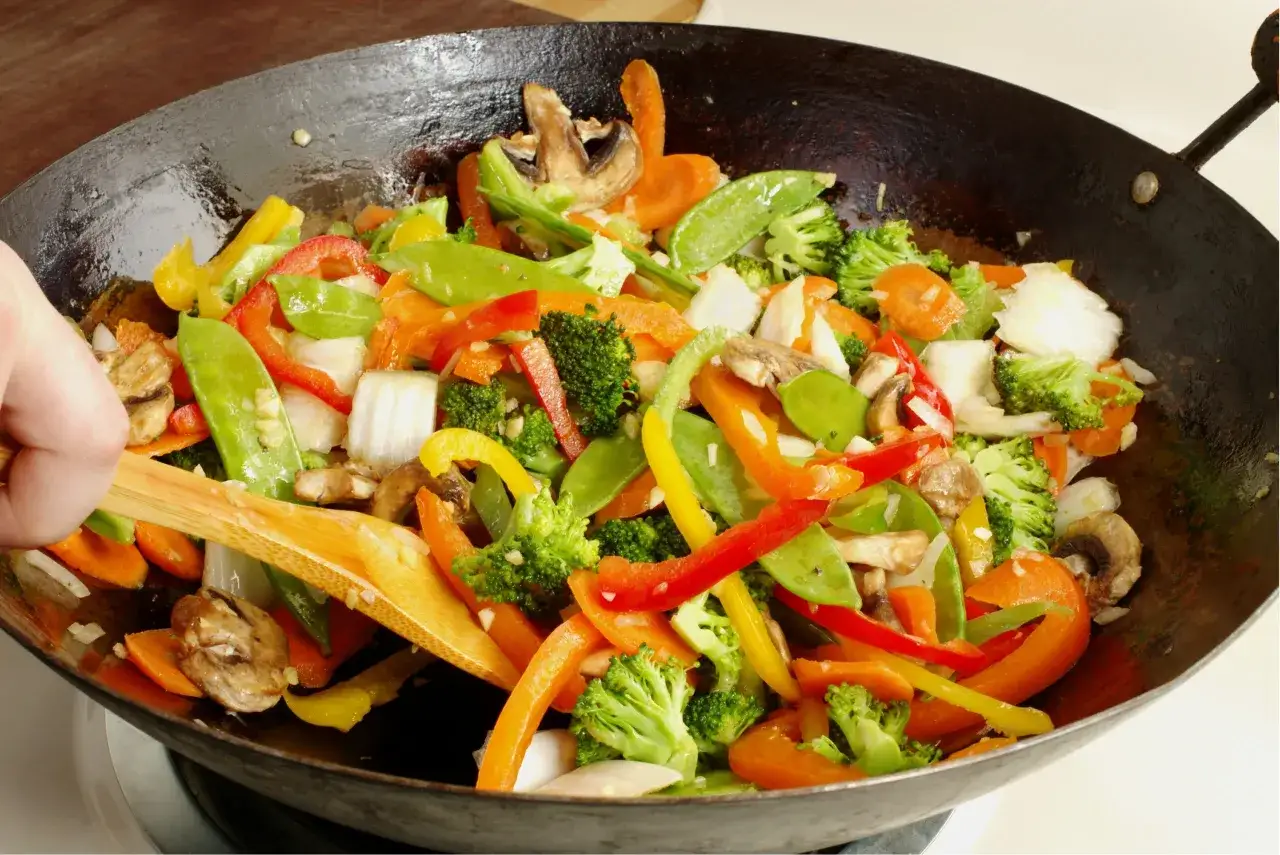 Un wok de verduras vibrante con br&oacute;coli, pimientos, zanahorias y guisantes, removido con una esp&aacute;tula de madera.