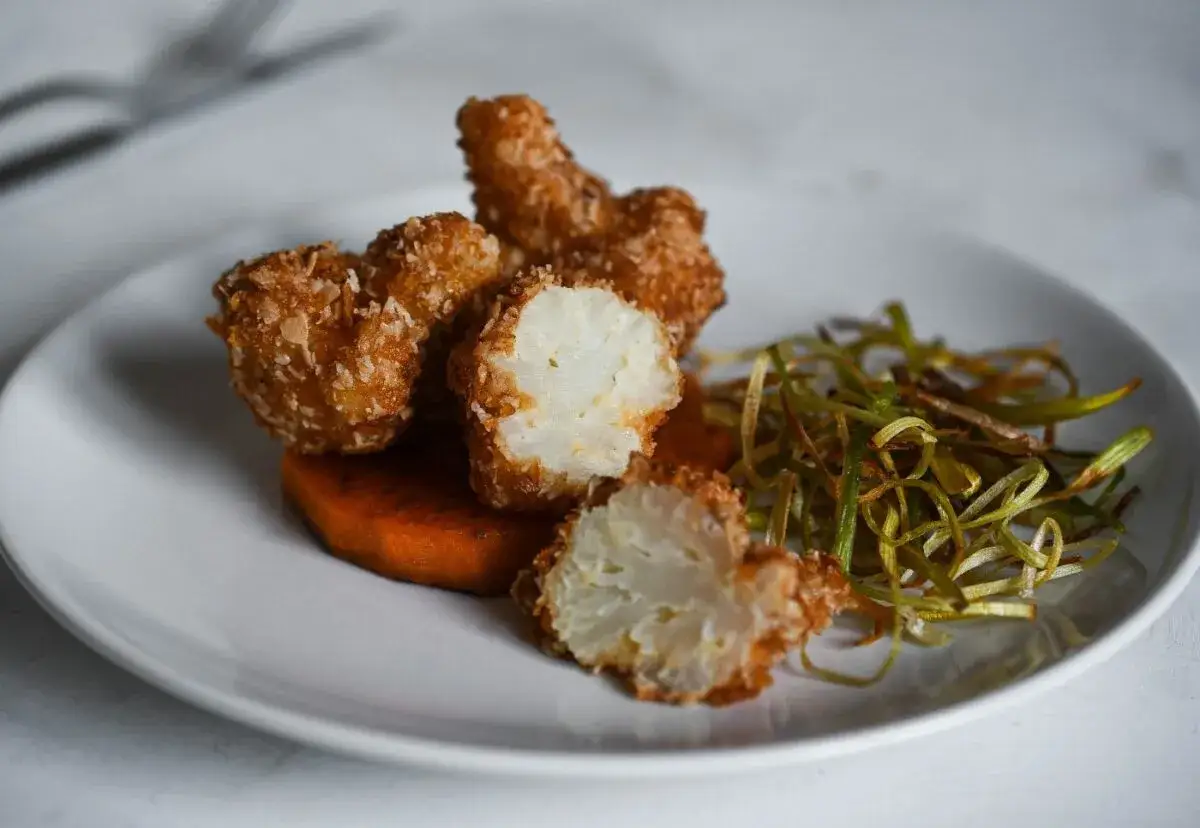 Crujiente coliflor rebozada con coco rallado, servida sobre puré de calabaza y acompañada de puerro crujiente.