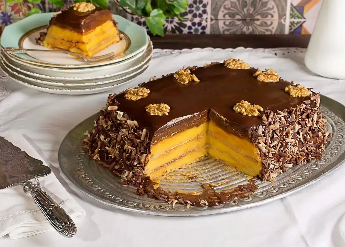 Deliciosa tarta de galletas y chocolate, con capas amarillas y cobertura brillante de chocolate, decorada con nueces y virutas de chocolate.