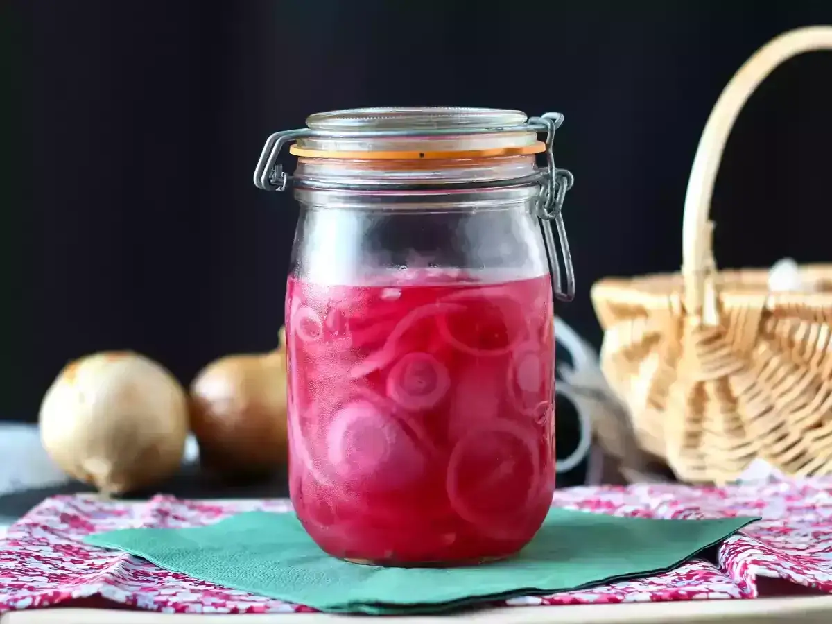 Frasco de cebolla encurtida con rodajas rosadas vibrantes. Dos cebollas crudas al lado, una cesta de mimbre al fondo.