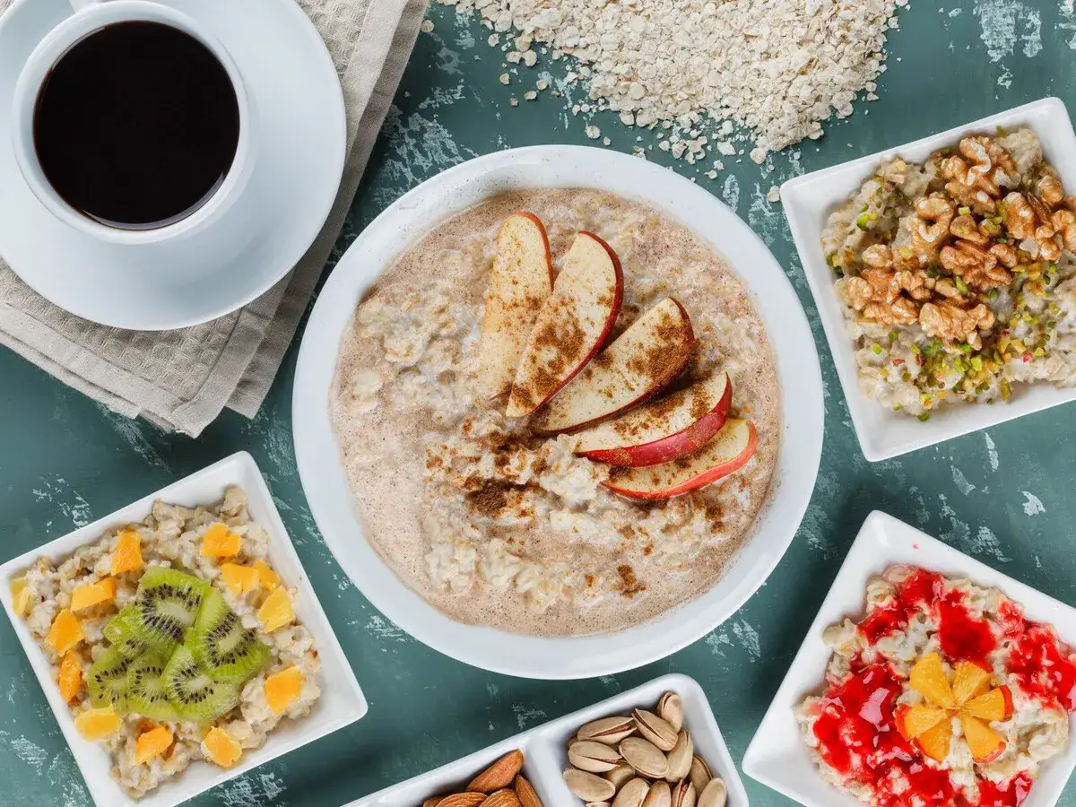Un delicioso desayuno con porridge de avena, café y frutas. El porridge principal tiene rodajas de manzana y canela. Otros cuencos muestran avena con kiwi, naranja, nueces, pistachos y mermelada.