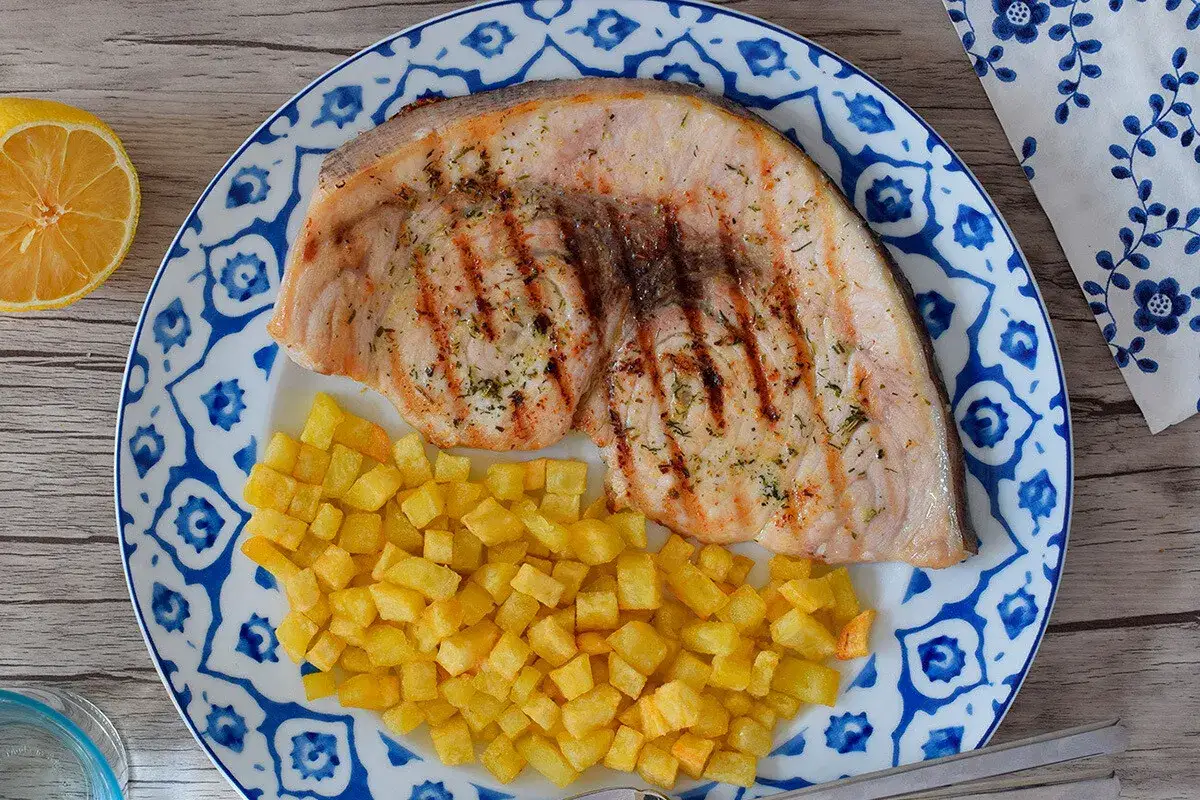 Delicioso emperador a la plancha con patatas fritas crujientes, servido en un plato con diseño azul y blanco.