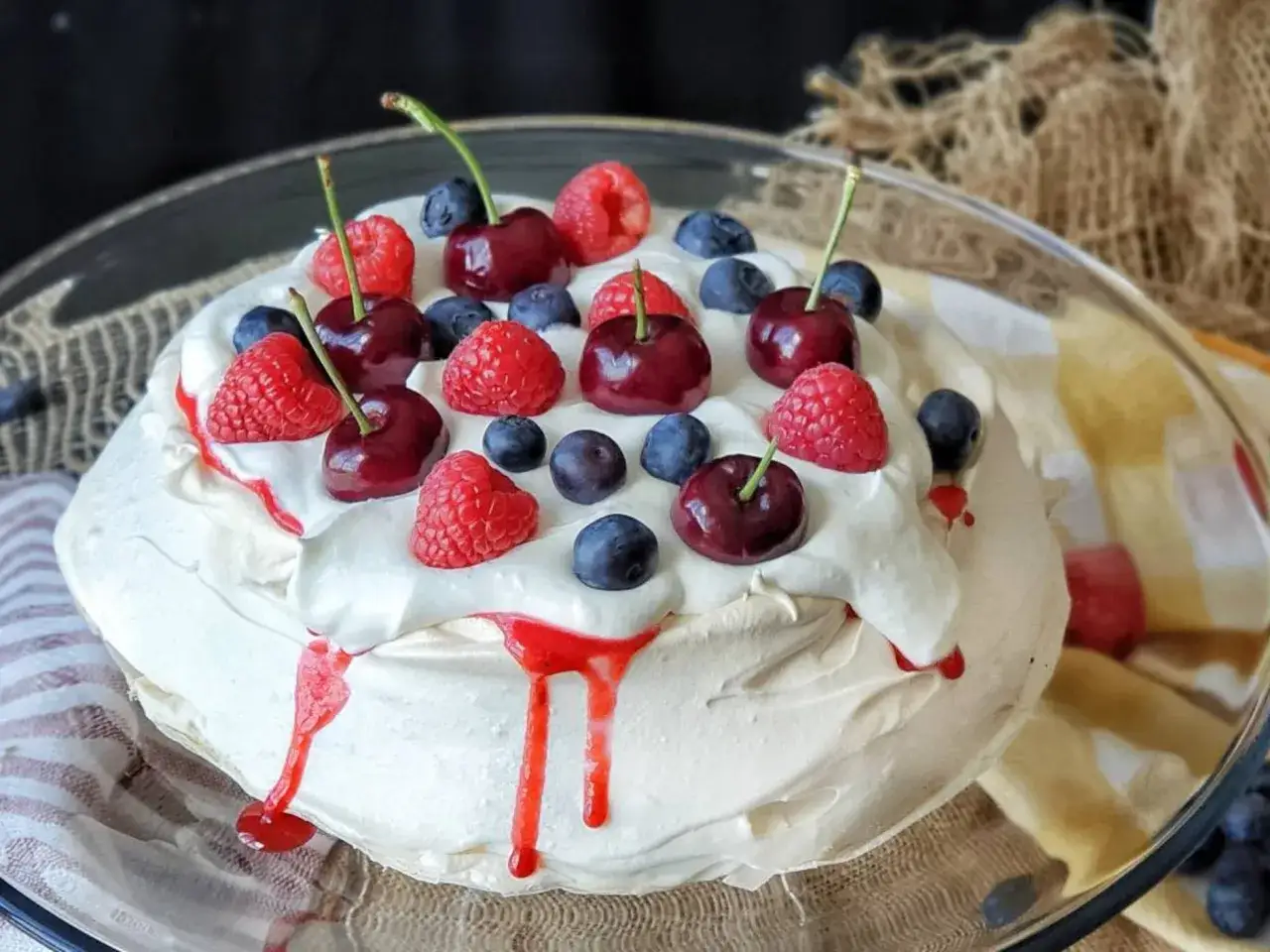 Tarta pavlova con crema batida, cerezas, frambuesas y ar&aacute;ndanos, decorada con un sirope rojo.