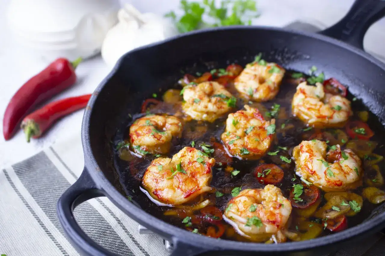 Gambas al pil pil chisporroteando en una sart&eacute;n de hierro fundido, con guindillas rojas y ajo al fondo.