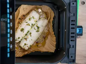 Dos filetes de merluza con hierbas cocinándose en papel de horno dentro de una freidora de aire.