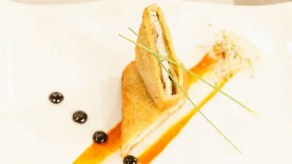 Un crujiente san jacobo, cortado por la mitad, reposa sobre una salsa naranja. Brotes y puntos oscuros decoran el plato.