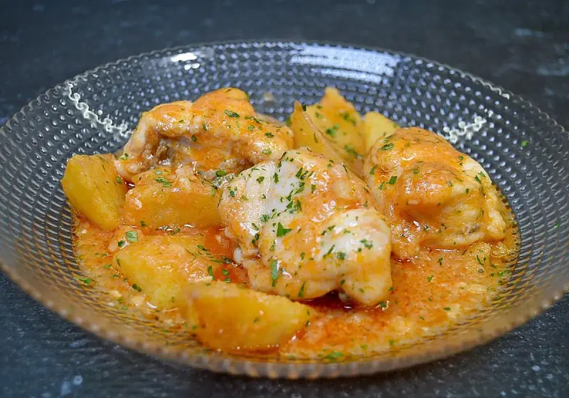 Rape a la marinera con patatas, servido en un cuenco de cristal con salsa naranja y perejil picado.
