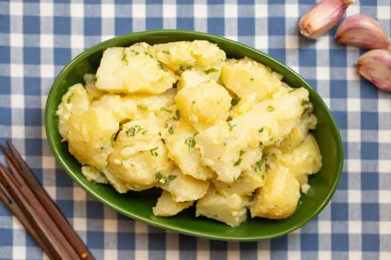 Un tazón verde lleno de patatas alioli cremosas, adornadas con perejil fresco. A su lado, ajos y tenedores de madera sobre un mantel de cuadros azules.