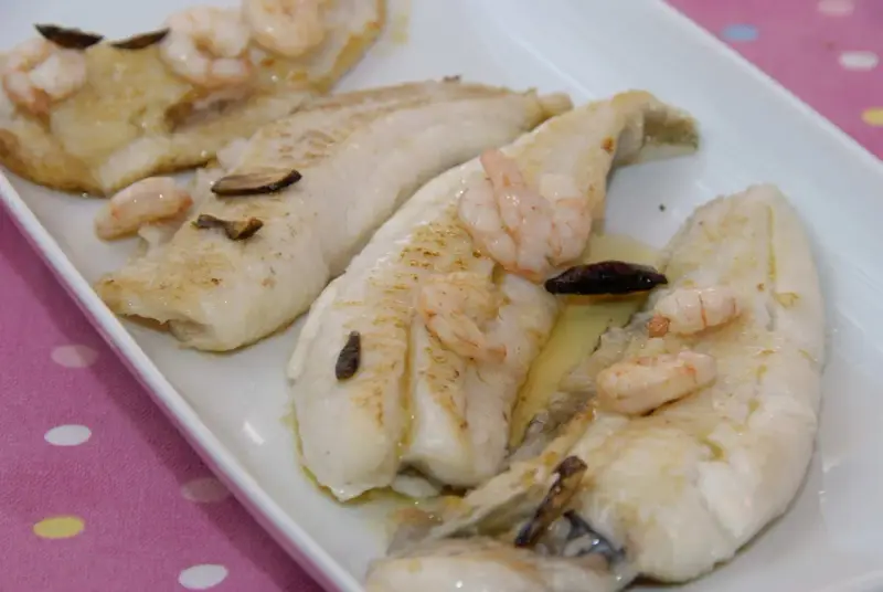 Filetes de gallo a la plancha con gambas y ajo laminado, servidos en una bandeja blanca.